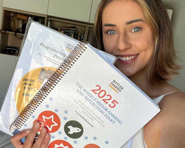 Woman holding a spiral-bound planner with 2025 programming planning on it in a kitchen.