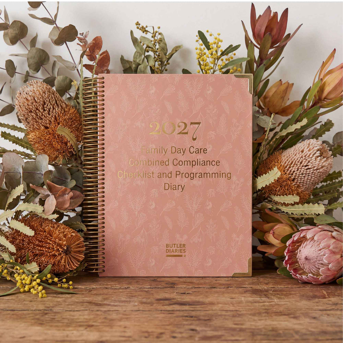 Pink diary titled '2027 Family Day Care Combined Compliance Checklist and Programming Diary' surrounded by flowers on a wooden surface.