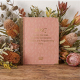 Pink diary titled '2027 Family Day Care Combined Compliance Checklist and Programming Diary' surrounded by flowers on a wooden surface.