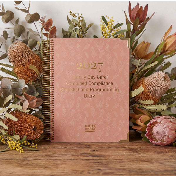 Pink diary titled '2027 Family Day Care Combined Compliance Checklist and Programming Diary' surrounded by flowers on a wooden surface.