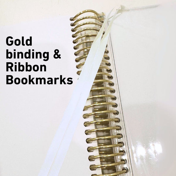 White notebook with gold binding and ribbon bookmarks on a white background