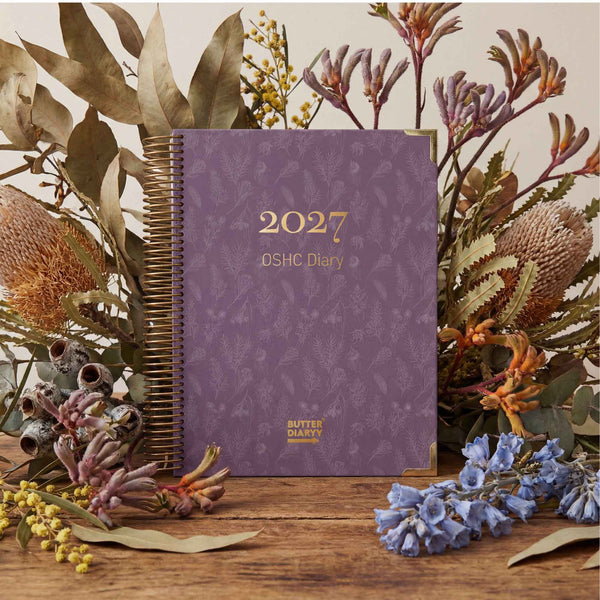 Purple diary with '2027 OSHC Diary' text, surrounded by floral and plant decorations on a wooden surface.