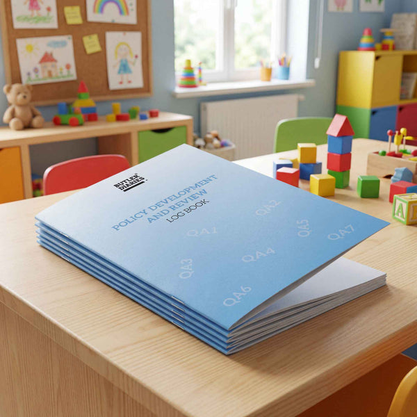 Stack of blue books titled 'Policy Development and Review Logbook' on a wooden table in a classroom setting with toys and children's artwork.