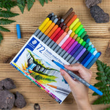 Set of Staedtler 72-color double-ended fibre-tips on a wooden surface with a hand holding a blue pen.