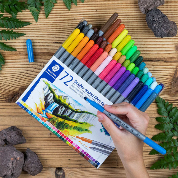 Set of Staedtler 72-color double-ended fibre-tips on a wooden surface with a hand holding a blue pen.