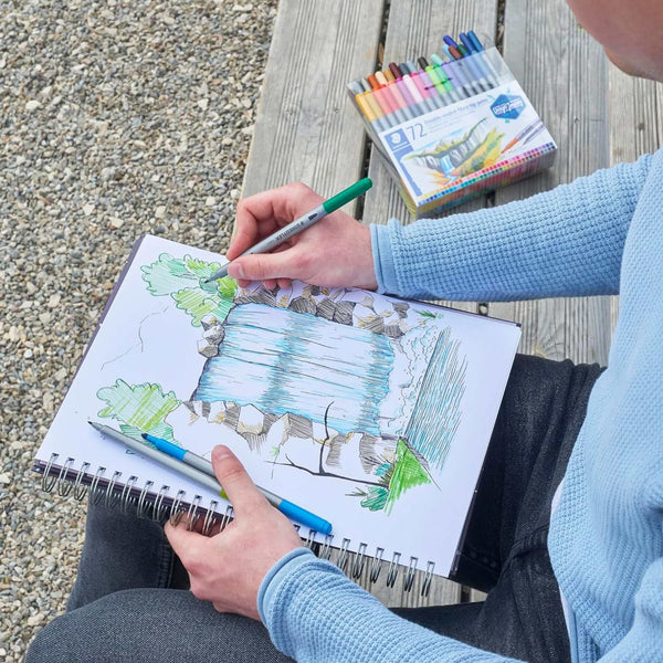 Person drawing in a sketchbook with colored markers on a wooden surface