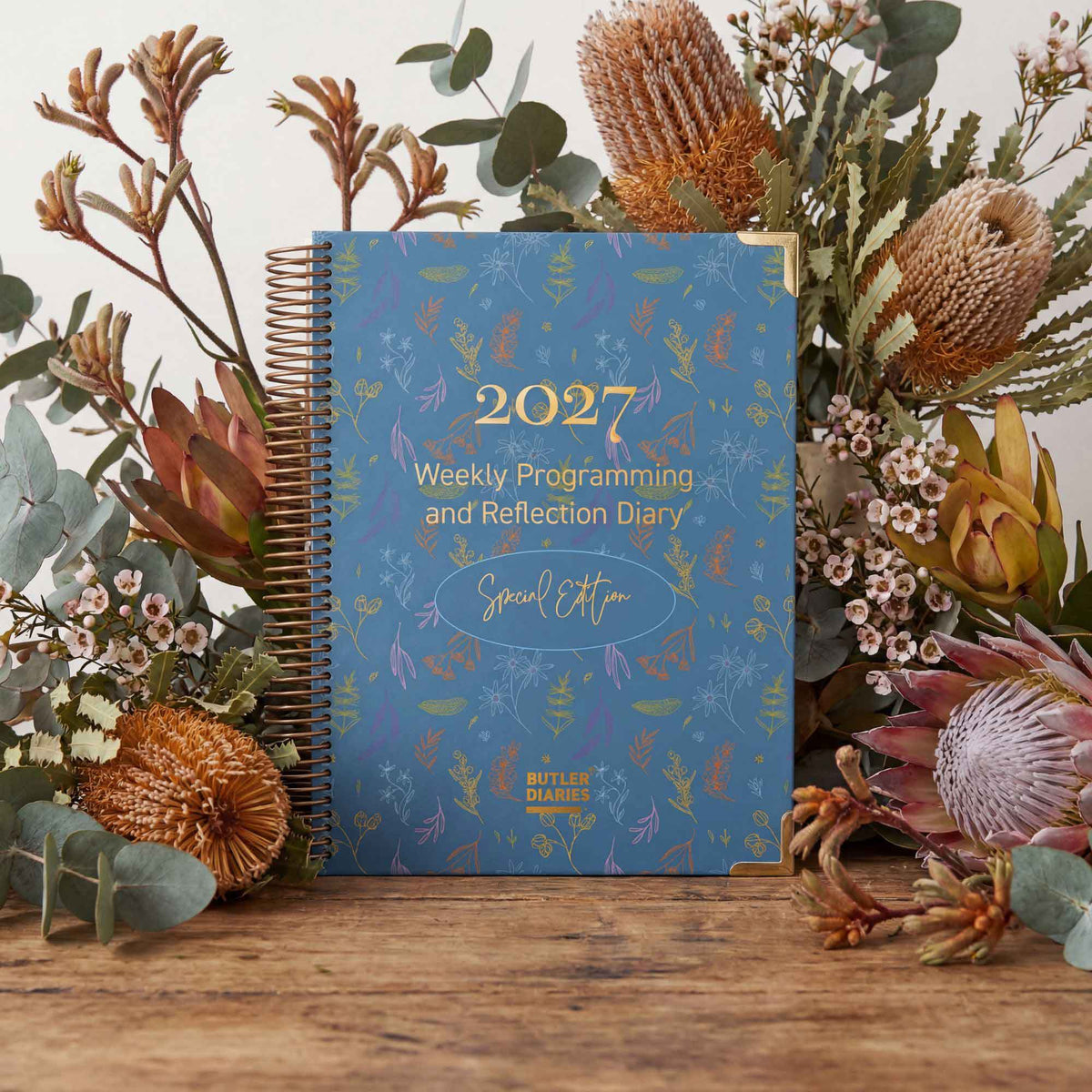 2027 weekly programming and reflection diary special edition with floral design on a wooden surface surrounded by flowers